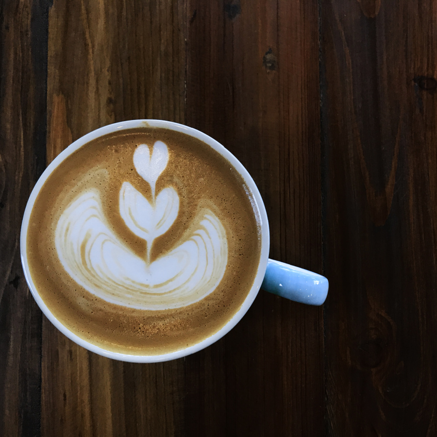 A wooden table with a cup of coffee with a heart growing out of a flower drawn in the foam.