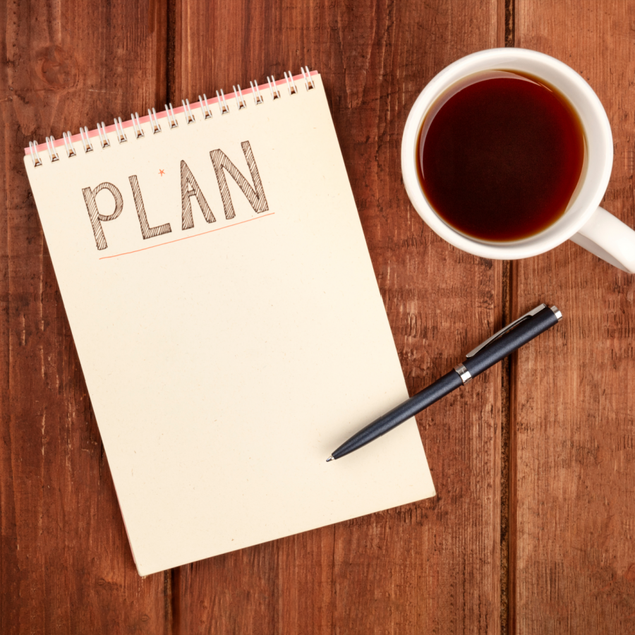 A notebook with “PLAN” on the top and a cup of coffee on top of a wooden table