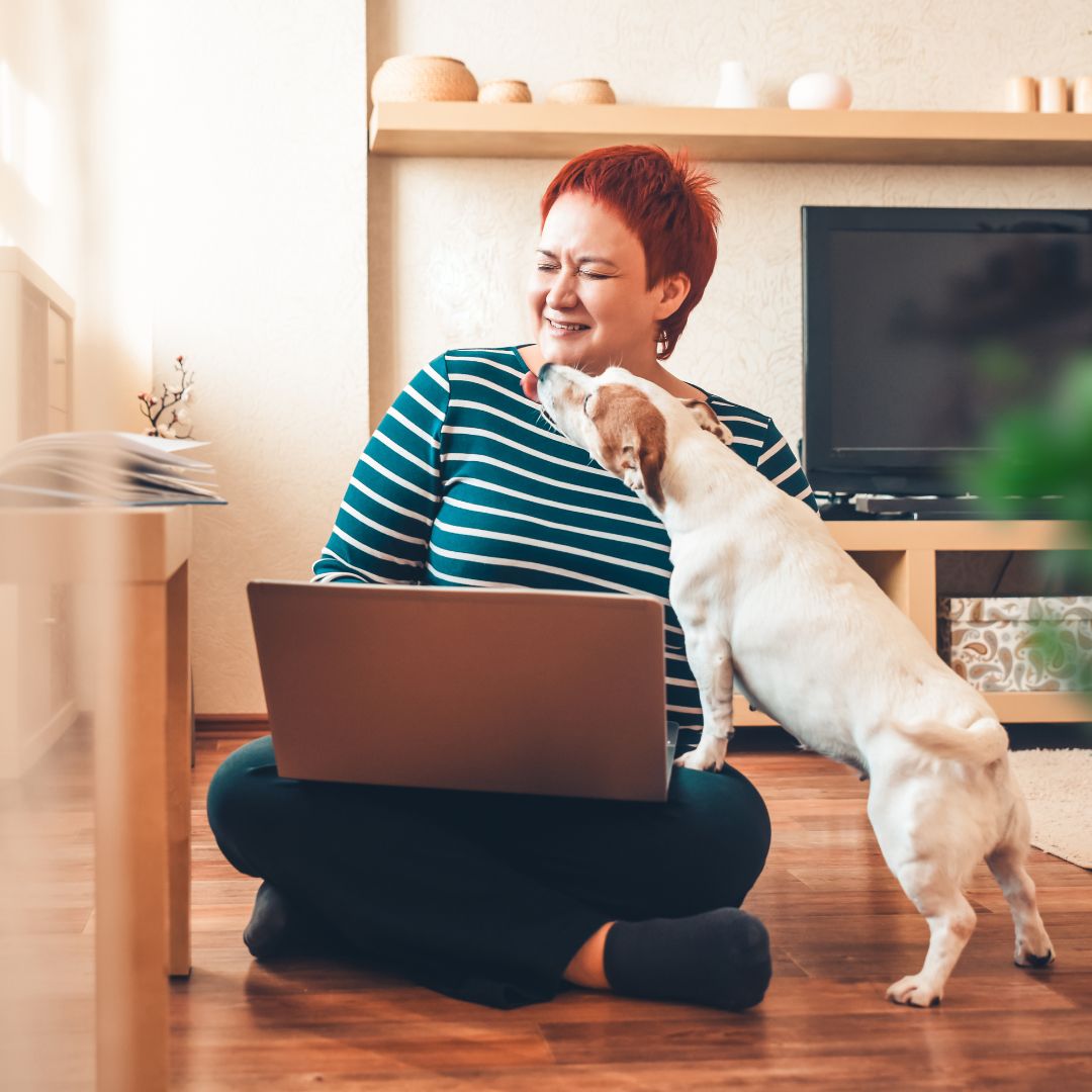 A woman sitting on the ground with a laptop open on her lap and a sweet dog stretching up to lick her chin.