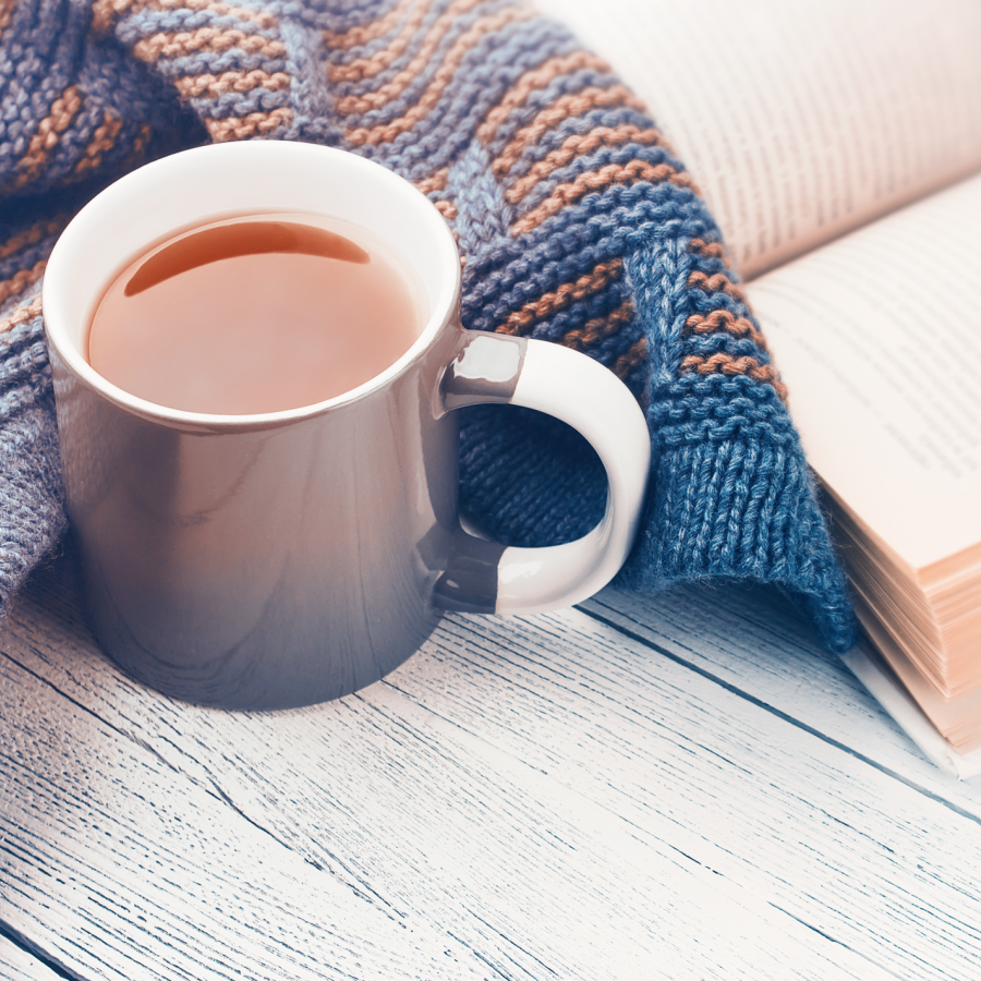 A full mug of coffee or tea resting on a wooden table next to a knit blanket and an open book