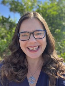 Jennifer Hicks, a white woman with glasses and long brown hair smiling, with green leaves in the background